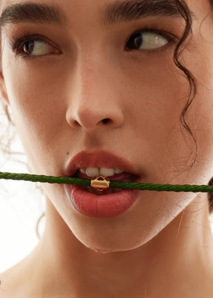 Modella morde delicatamente il cordino intrecciato verde in pelle con dettaglio dorato, Bracciale Siklah