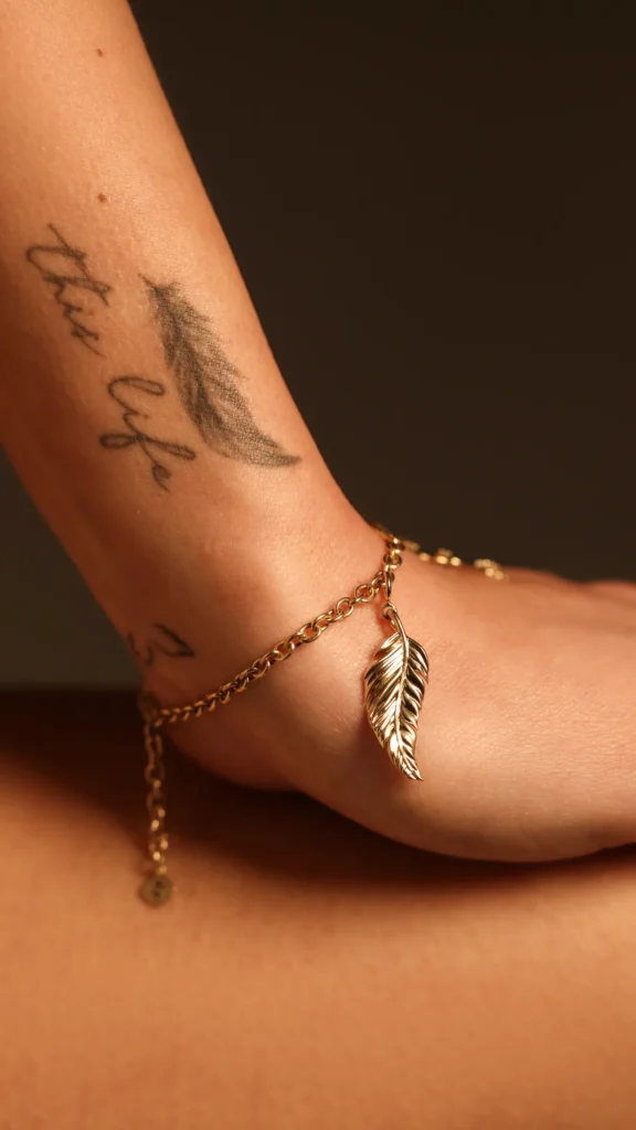 Charm Libertà Siklah a piuma in oro giallo, indossato su cavigliera con tatuaggio in vista
